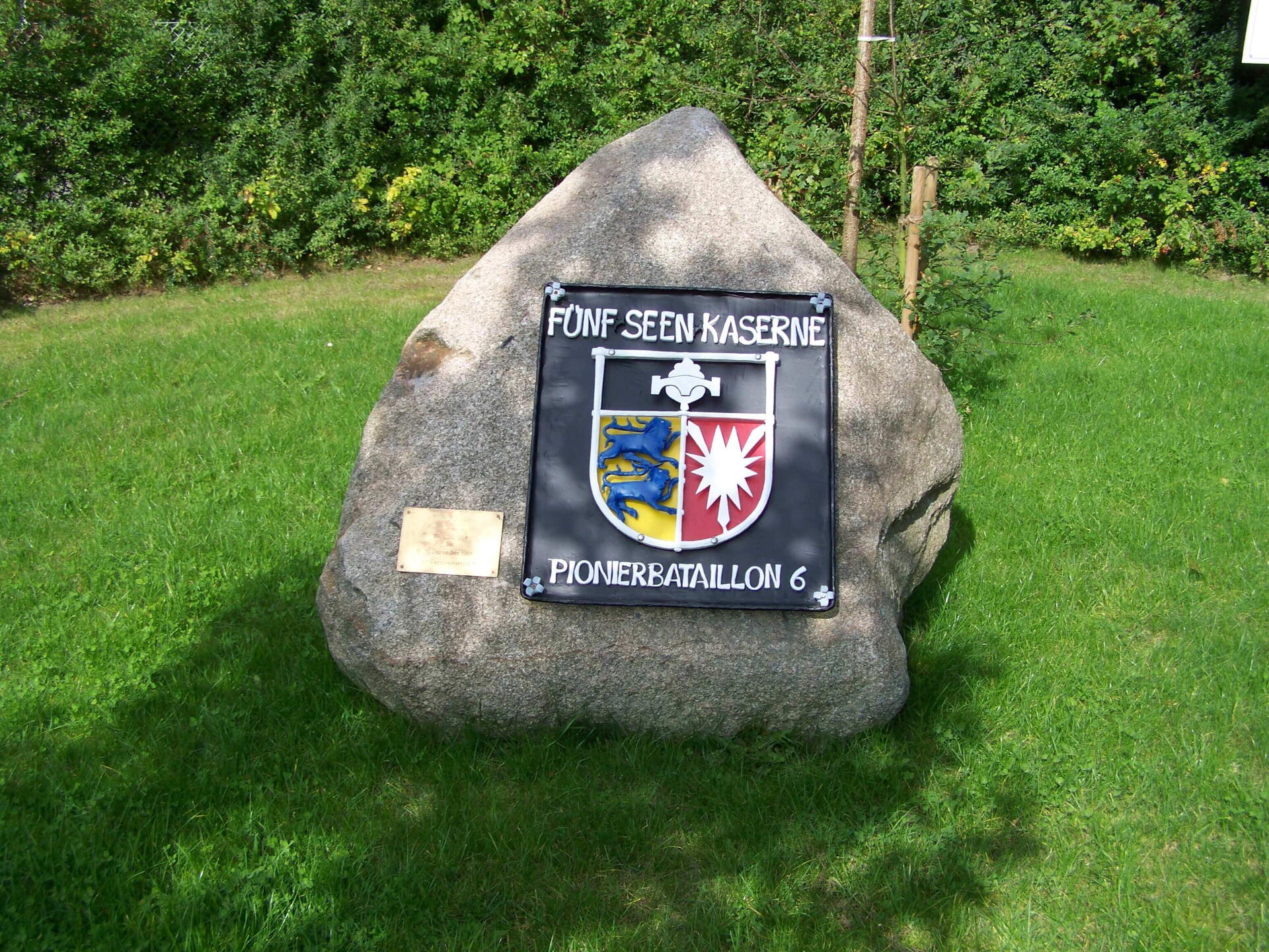 Gedenkstein mit Schild der Fünf-Seen-Kaserne, Pionierbataillon 6, auf grüner Wiese vor Bäumen.
