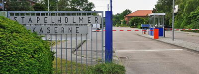 Eingang der Stapelholmer Kaserne mit Schranke und Wachhäuschen, umgeben von grüner Vegetation und einem klaren Himmel.