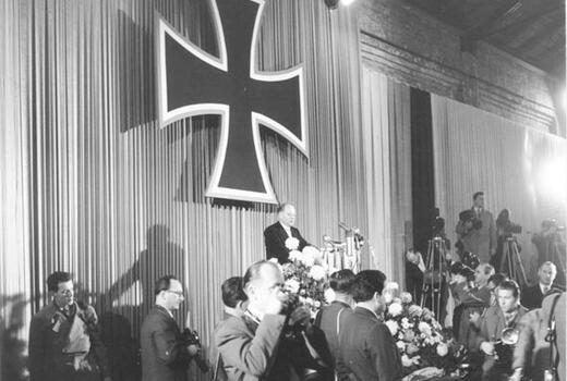 Ein Redner steht auf einer Bühne unter einem großen Kreuz, während Fotografen und Gäste die Ansprache verfolgen.
