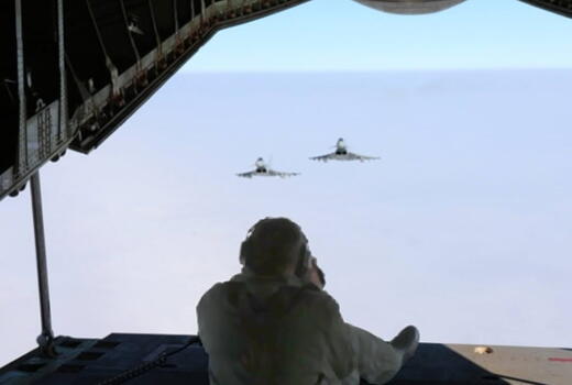 Ein Soldat sitzt im geöffneten Heck eines Flugzeugs und beobachtet zwei Kampfjets am Himmel.