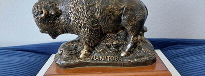 Eine bronzene Büffelstatue mit der Aufschrift "MANITOBA" steht auf einem Holzsockel.