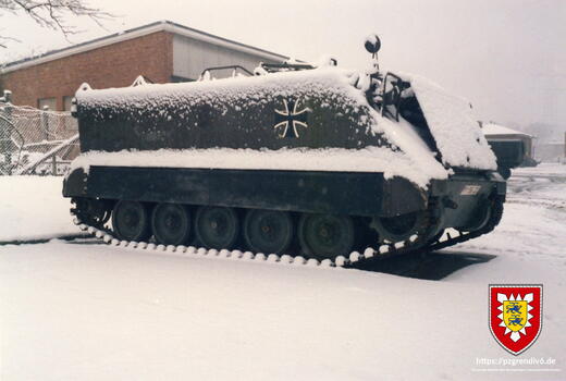 Ein schneebedeckter gepanzerter Truppentransporter steht auf einem verschneiten Gelände vor einem Gebäude.
