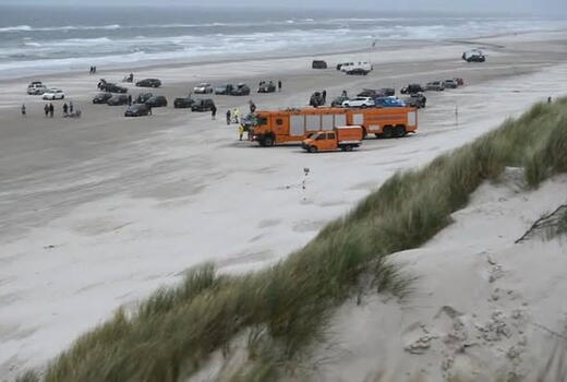Eine orangefarbene Rettungsfahrzeugflotte versammelt sich am stürmischen Strand, umgeben von mehreren parkenden Autos.
